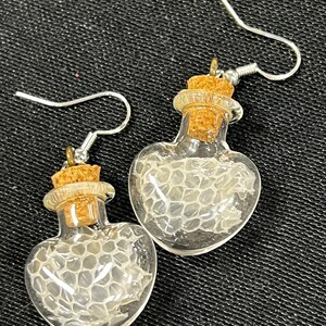 May include: A pair of heart-shaped glass bottle earrings with cork stoppers. Each bottle contains a piece of white snake skin.