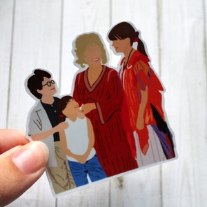 May include: A sticker featuring a group of four people, two adults and two children. The adults are wearing red and purple clothing, while the children are wearing white and blue clothing. The sticker is on a white wooden background.