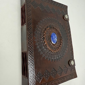 Könnte beinhalten: Ein dunkelbraunes Lederjournal mit geprägtem geometrischem Muster und einem zentralen, ovalen blauen Steineinsatz. Der Einband wird mit zwei Metalldruckknöpfen auf der rechten Seite verschlossen, und der Buchrücken ist mit sichtbarer Lederkordel gebunden.