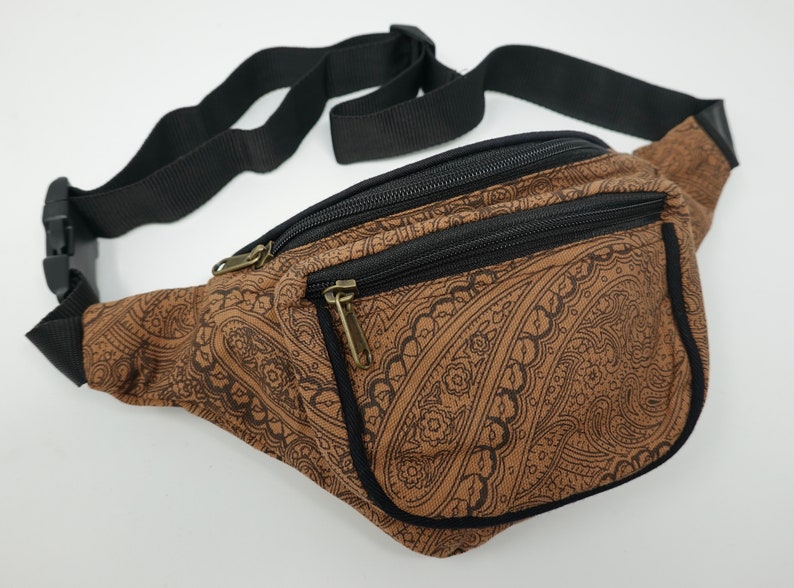 Op de afbeelding: Een bruine heuptas met een zwart paisley-patroon en een zwarte verstelbare riem. De tas heeft twee vakken met rits.