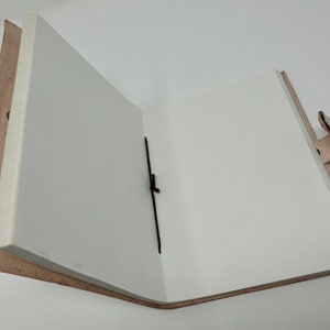 Puede incluir: Un diario de cuero marr&oacute;n abierto con p&aacute;ginas blancas en blanco y un marcador de cinta negro. El diario tiene un cierre de correa de cuero.