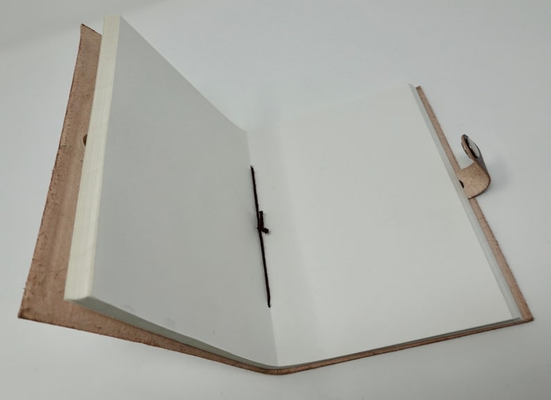 Puede incluir: Un diario de cuero marr&oacute;n abierto con p&aacute;ginas blancas en blanco y un marcador de cinta negro. El diario tiene un cierre de correa de cuero.