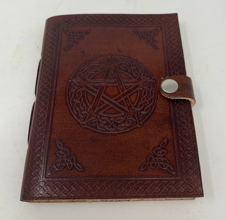 May include: A brown leather journal with a pentagonal star design embossed on the front cover. The journal has a strap closure with a metal button.