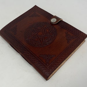 May include: A brown leather journal with a pentagonal star design embossed on the front cover. The journal has a leather strap closure with a metal button.