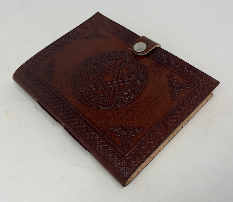 May include: A brown leather journal with a pentagonal star design embossed on the front cover. The journal has a leather strap closure with a metal button.