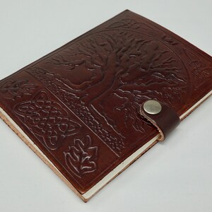 Puede incluir: Un diario de cuero marr&oacute;n con un dise&ntilde;o de &aacute;rbol de la vida y un borde de nudo celta. El diario tiene un cierre de correa de cuero con un bot&oacute;n de metal.