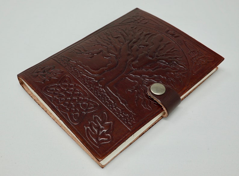 Puede incluir: Un diario de cuero marr&oacute;n con un dise&ntilde;o de &aacute;rbol de la vida y un borde de nudo celta. El diario tiene un cierre de correa de cuero con un bot&oacute;n de metal.