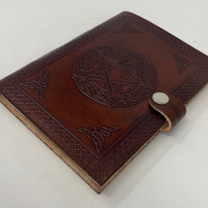 May include: A brown leather journal with a pen loop and a silver button closure. The cover features an embossed Celtic knot design with a pentagram in the center.