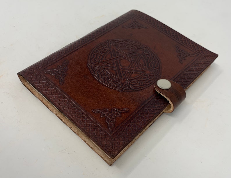 May include: A brown leather journal with a pen loop and a silver button closure. The cover features an embossed Celtic knot design with a pentagram in the center.