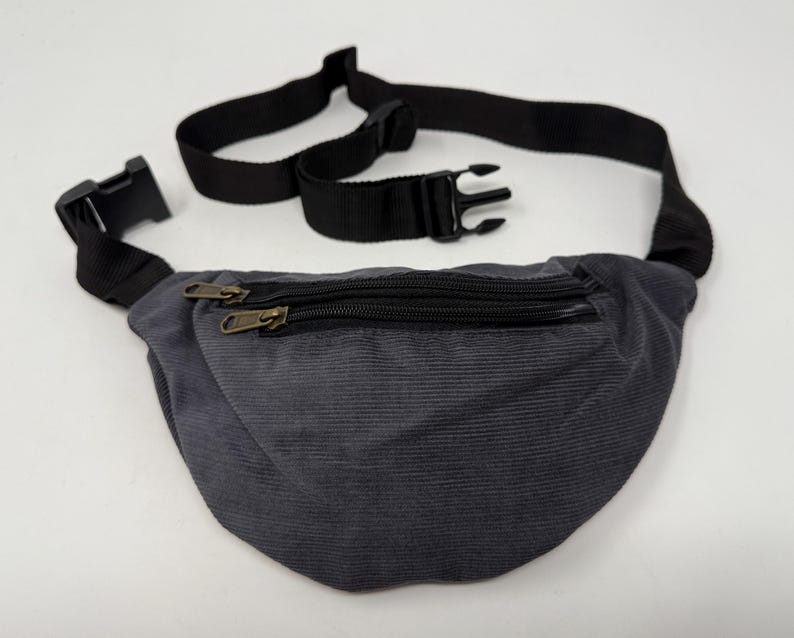May include: A gray corduroy fanny pack with a black adjustable strap and two zippered compartments.