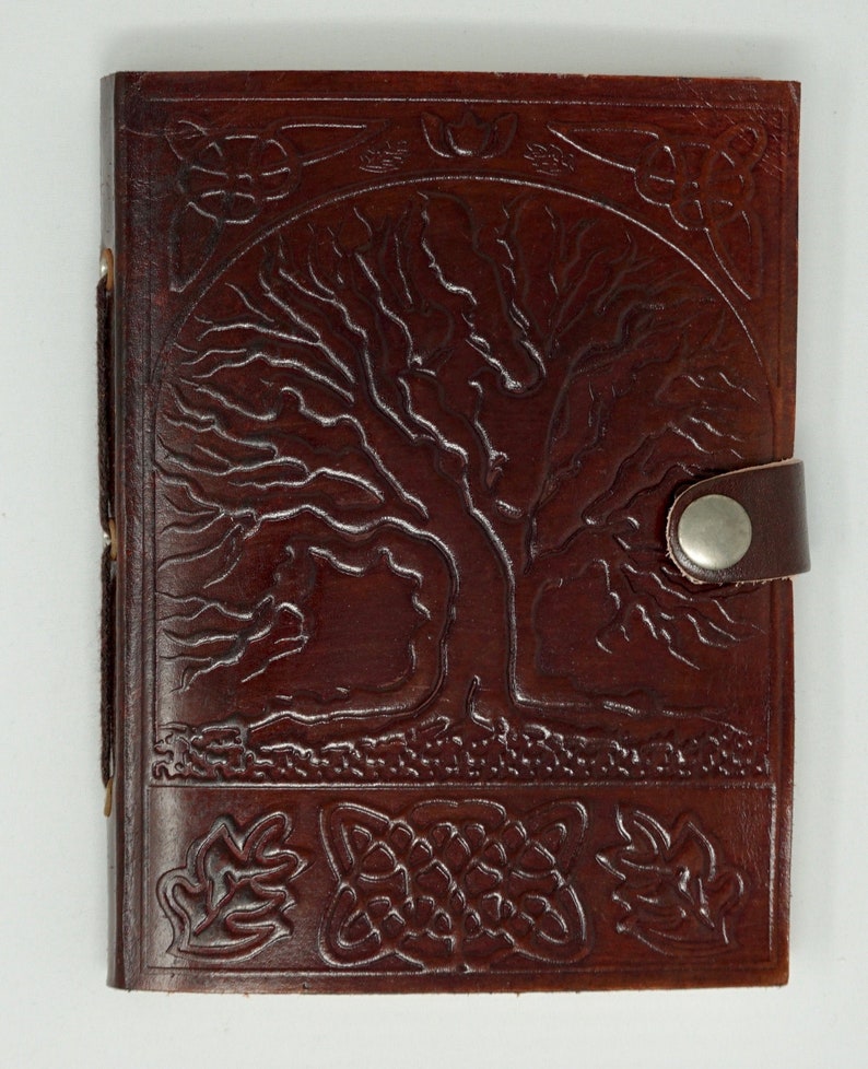 Puede incluir: Un diario de cuero marr&oacute;n con un dise&ntilde;o de &aacute;rbol de la vida en relieve en la portada. El diario tiene un cierre de bot&oacute;n met&aacute;lico y una correa decorativa.