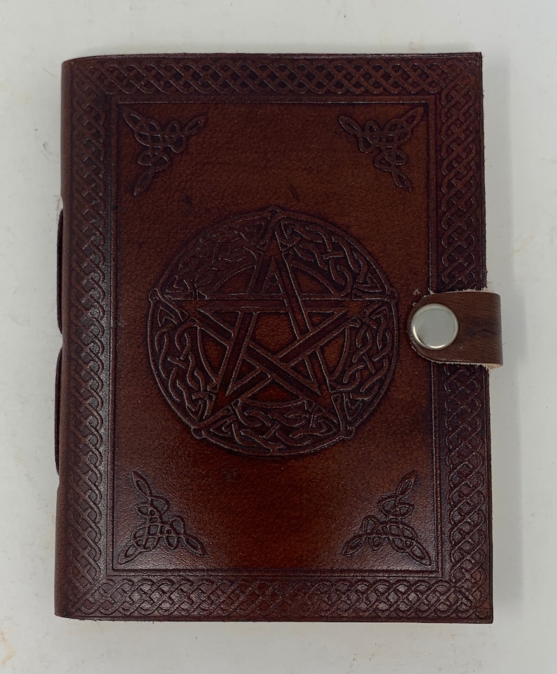May include: A brown leather journal with a pentacle design embossed on the front cover. The journal has a decorative border and a leather strap with a metal button closure.