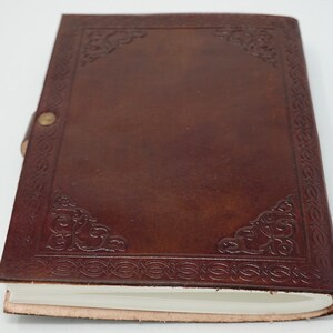 Puede incluir: Un diario de cuero marr&oacute;n con un dise&ntilde;o decorativo en relieve alrededor de los bordes. El diario tiene una p&aacute;gina blanca en blanco visible.