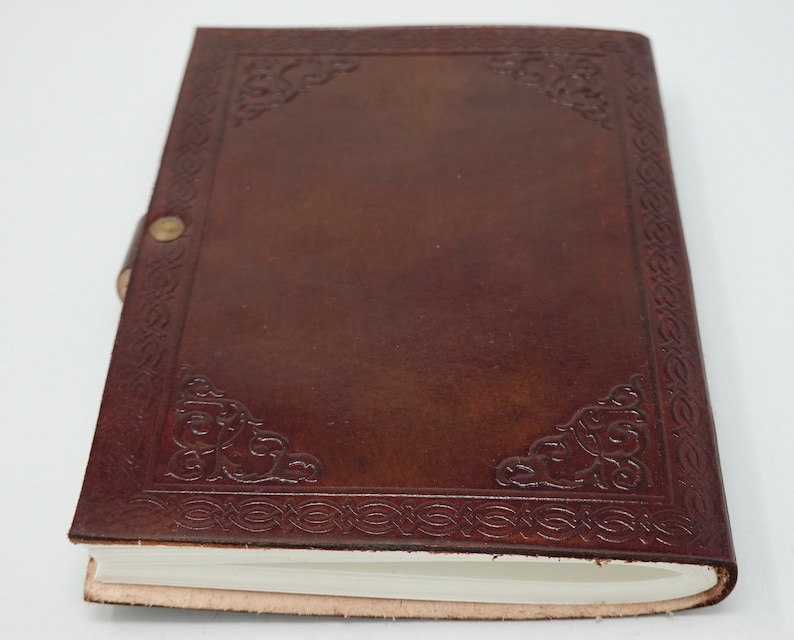 Puede incluir: Un diario de cuero marr&oacute;n con un dise&ntilde;o decorativo en relieve alrededor de los bordes. El diario tiene una p&aacute;gina blanca en blanco visible.