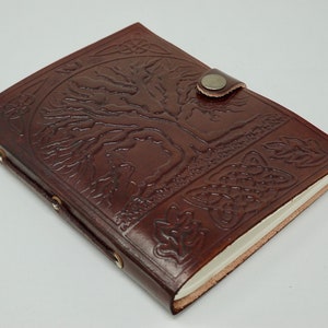 Puede incluir: Un diario de cuero marr&oacute;n con un dise&ntilde;o de &aacute;rbol de la vida en relieve en la portada. El diario tiene un cierre de correa y un interior de papel blanco.