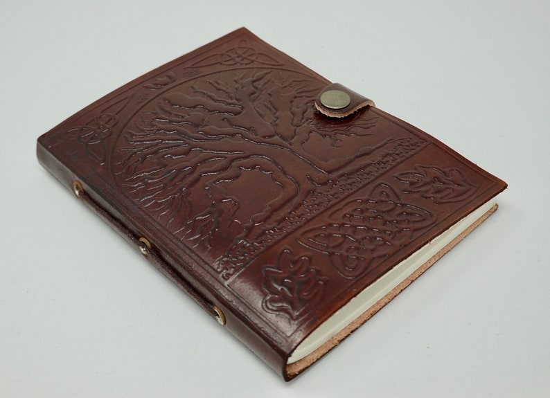 Puede incluir: Un diario de cuero marr&oacute;n con un dise&ntilde;o de &aacute;rbol de la vida en relieve en la portada. El diario tiene un cierre de correa y un interior de papel blanco.