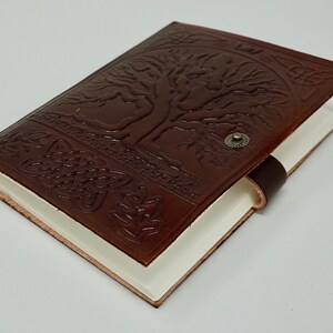 Puede incluir: Un diario de cuero marr&oacute;n con un dise&ntilde;o de &aacute;rbol de la vida en relieve en la portada. El diario tiene un cierre de correa de cuero y un bot&oacute;n de metal.