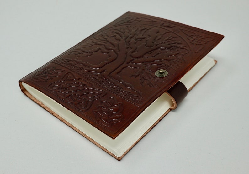 Puede incluir: Un diario de cuero marr&oacute;n con un dise&ntilde;o de &aacute;rbol de la vida en relieve en la portada. El diario tiene un cierre de correa de cuero y un bot&oacute;n de metal.