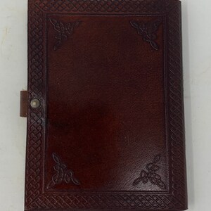 May include: A brown leather journal with a Celtic knot design embossed on the cover. The journal has a flap closure and a metal button.