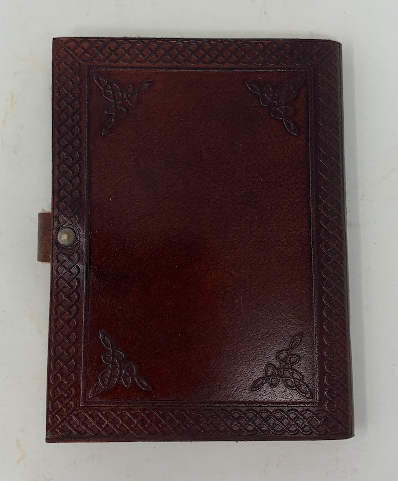May include: A brown leather journal with a Celtic knot design embossed on the cover. The journal has a flap closure and a metal button.