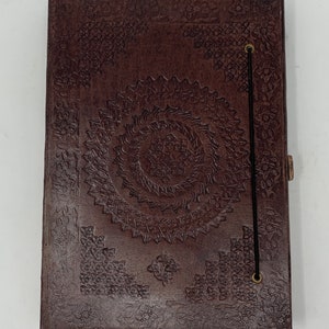 May include: A dark brown leather journal with an embossed floral and spiral design. The cover features a central spiral pattern surrounded by floral corner accents. A black elastic band and a small clasp secure the journal.