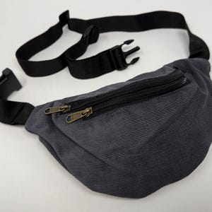 May include: A gray corduroy fanny pack with a black adjustable strap and two zipper closures.