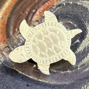 May include: A detailed, cream-colored turtle figurine with etched shell patterns and red eye accents. The figurine is set against a textured, multi-toned background, showcasing its intricate design and craftsmanship.