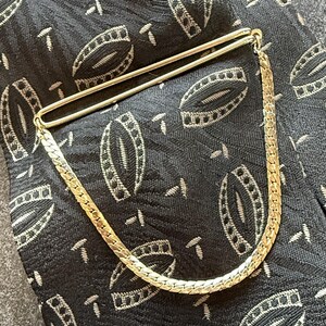 May include: A gold-toned tie bar featuring a sleek, polished metal clip attached to a flat, braided curb chain. The accessory is positioned against a black fabric background that displays a repeating pattern of small, light-colored oval shapes.