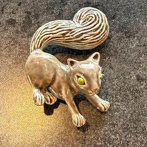 May include: A ceramic cat figurine in a crouching pose. The cat is gray with green eyes and a large, curled tail with brown and white stripes. The figurine is on a dark, textured surface.