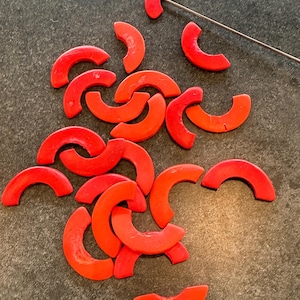 May include: A collection of bright red, semi-circular objects scattered on a dark gray surface. The objects are evenly colored and appear to be made of a solid material. A thin, silver metal rod is also present, possibly for manipulation or display.