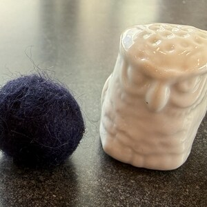 May include: A navy blue wool ball next to a white ceramic owl thimble. The thimble has a detailed owl design with textured feathers and a rounded top. The wool ball has a slightly fuzzy texture.