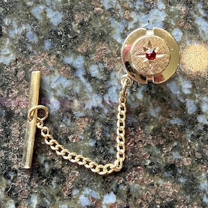 May include: A gold-toned tie tack with a circular design featuring a red gemstone at the center. The tack is connected to a gold chain and a bar. The background is a dark, speckled surface.