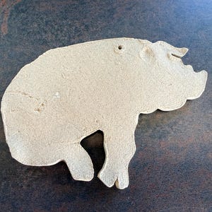 May include: A beige, ceramic pig-shaped ornament with a hole for hanging. The pig is depicted in profile, with visible details like the snout, ear, and legs. The ornament has a textured surface and is likely handmade.
