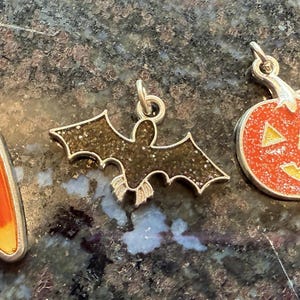 May include: Three Halloween-themed charms. One is a silver-toned, triangular charm with orange and yellow enamel. The second is a bat-shaped charm with glitter. The third is a pumpkin charm with a jack-o'-lantern face.