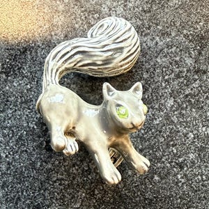 May include: A gray squirrel brooch with a curled tail and green eyes. The brooch is made of metal and has a detailed design. The squirrel is in a running pose, with its paws extended. The tail is striped with white and gray.