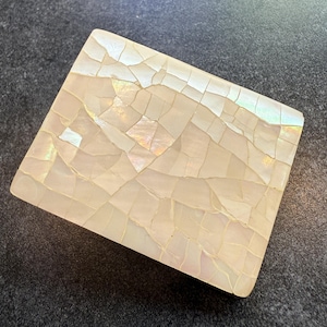May include: A square, iridescent, mother-of-pearl box. The surface is made of small, irregular pieces, creating a mosaic effect. The box has a pearlescent sheen, reflecting light with hints of pink, green, and gold. The box is set against a dark gray background.