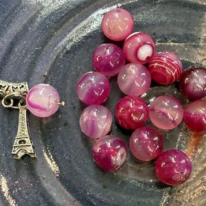 May include: A collection of pink and purple agate beads with a silver Eiffel Tower charm. The beads vary in color and pattern, with some having a striped design. The charm is detailed with intricate carvings.