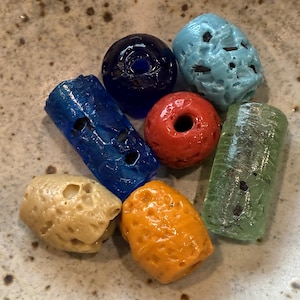 May include: A collection of colorful, textured glass beads in various shapes. The beads include shades of blue, red, green, yellow, and orange. Some beads are cylindrical, while others are round or irregular. The beads have holes for stringing.