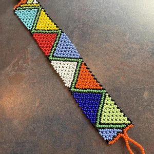 May include: A beaded bracelet with a geometric design. The bracelet features triangles in various colors, including blue, yellow, red, white, orange, and green, outlined in black. The bracelet has an orange loop at one end and an orange bead at the other.