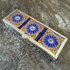 May include: A rectangular box featuring three square tiles on the lid. Each tile displays a central blue and light purple circular mandala pattern surrounded by a yellow and orange rope-like border. The box has a metallic frame with a dark cutout design.