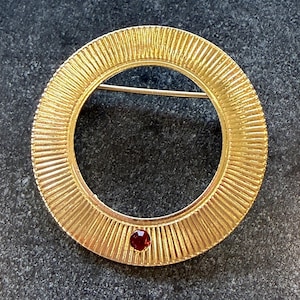 May include: A gold-toned circular brooch with a textured, ridged design. The brooch features a small, round, red gemstone at the bottom. The pin is a classic jewelry piece.