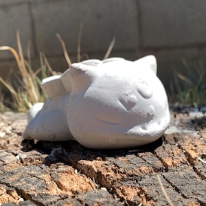 May include: A small, white, concrete sculpture of a cartoon cat lying down. The cat has a smiling face and is resting on a weathered, brown tree stump. The background features a gray brick wall and dry grass.