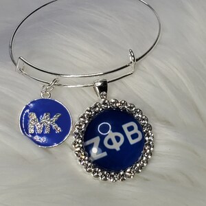 Puede incluir: Un brazalete de plata con dos dijes. Un dije es azul con letras blancas que dicen "MK" y el otro dije es azul con letras blancas que dicen "ZΦB". El dije con las letras "ZΦB" está rodeado por un anillo plateado de diamantes de imitación.