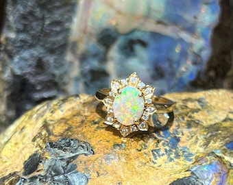 Op de afbeelding: Een gouden ring met een witte opaal-edelsteen omringd door een halo van kleine diamanten. De ring ligt op een rots met een kleurrijke achtergrond.