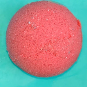 May include: A round, red bath bomb with a textured surface and subtle glitter accents. The bath bomb is set against a turquoise background, creating a vibrant contrast. This product is designed for use in the bath.