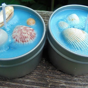 May include: Two round, silver-colored tin candles with blue wax, decorated with seashells. The candles have white wicks and glitter accents, creating a beach or ocean theme. The seashells vary in size and color.