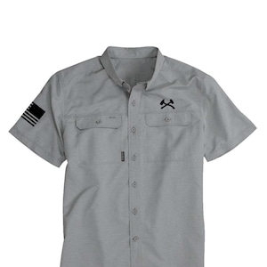 May include: A light gray short-sleeved shirt with two chest pockets and a button-down front. The shirt features a black graphic of crossed axes on the right chest and a black flag design on the left sleeve. The shirt has a classic collar.
