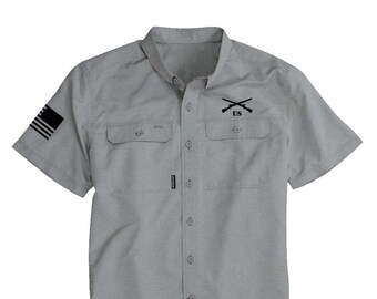 Puede incluir: Una camisa gris de manga corta con botones, con una bandera estadounidense en blanco y negro en la manga izquierda y un diseño bordado en negro de rifles cruzados con "US" encima en el bolsillo del pecho derecho.