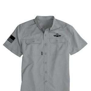 May include: A gray short-sleeved button-down shirt with a black and white American flag on the left sleeve and a black logo on the right chest pocket.