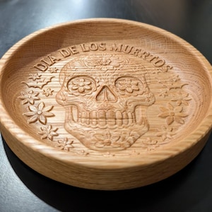 Peut inclure: Un bol rond en bois clair avec un motif de crâne en sucre sculpté. Les mots "DIA DE LOS MUERTOS" sont gravés sur le bord. Le bol est de couleur marron clair et présente des motifs floraux.